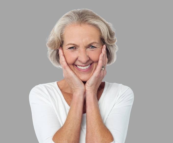 Smiling mature woman with hands to face in white shirt on gray background.